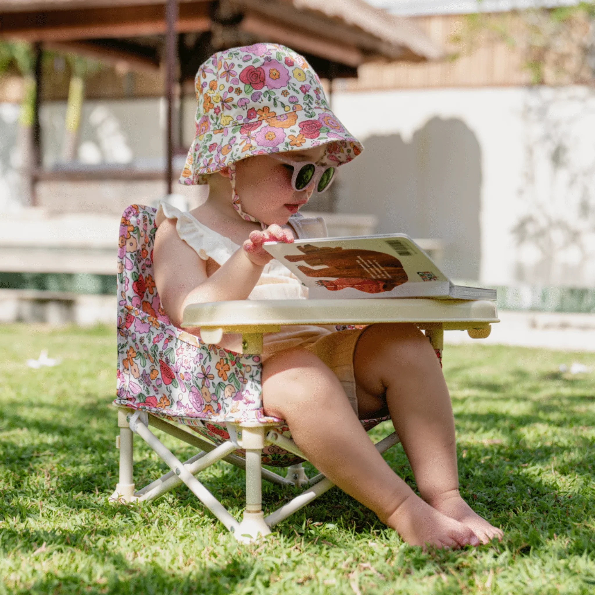 Toddler in a sun hat reading a book in a Paloma baby chair on the grass