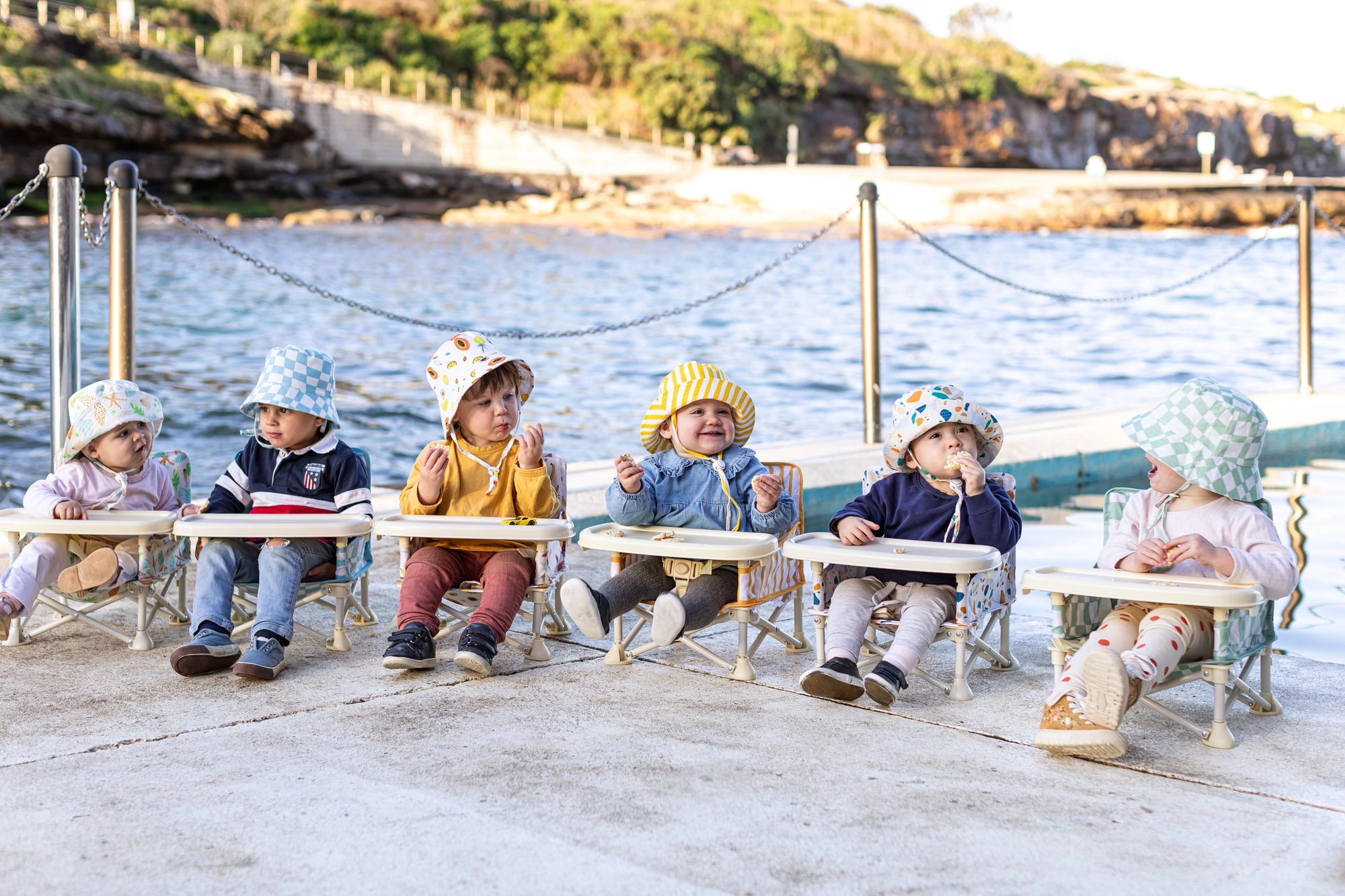 Six babies in IZIMINI chairs and sun hats lined up along a waterside jetty