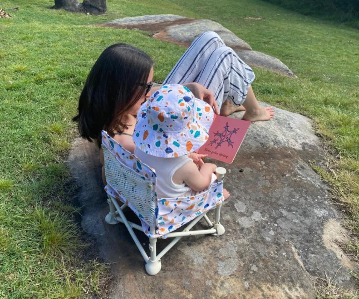 Mum reading to a baby in a camping chair and floral hat on a rock