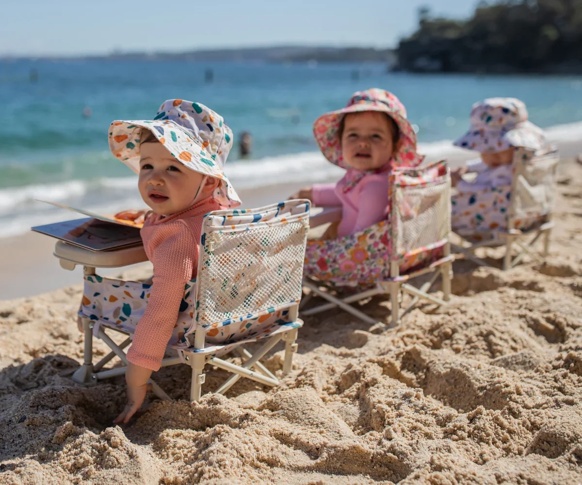 Three babies in camping chairs and matching hats on the sand
