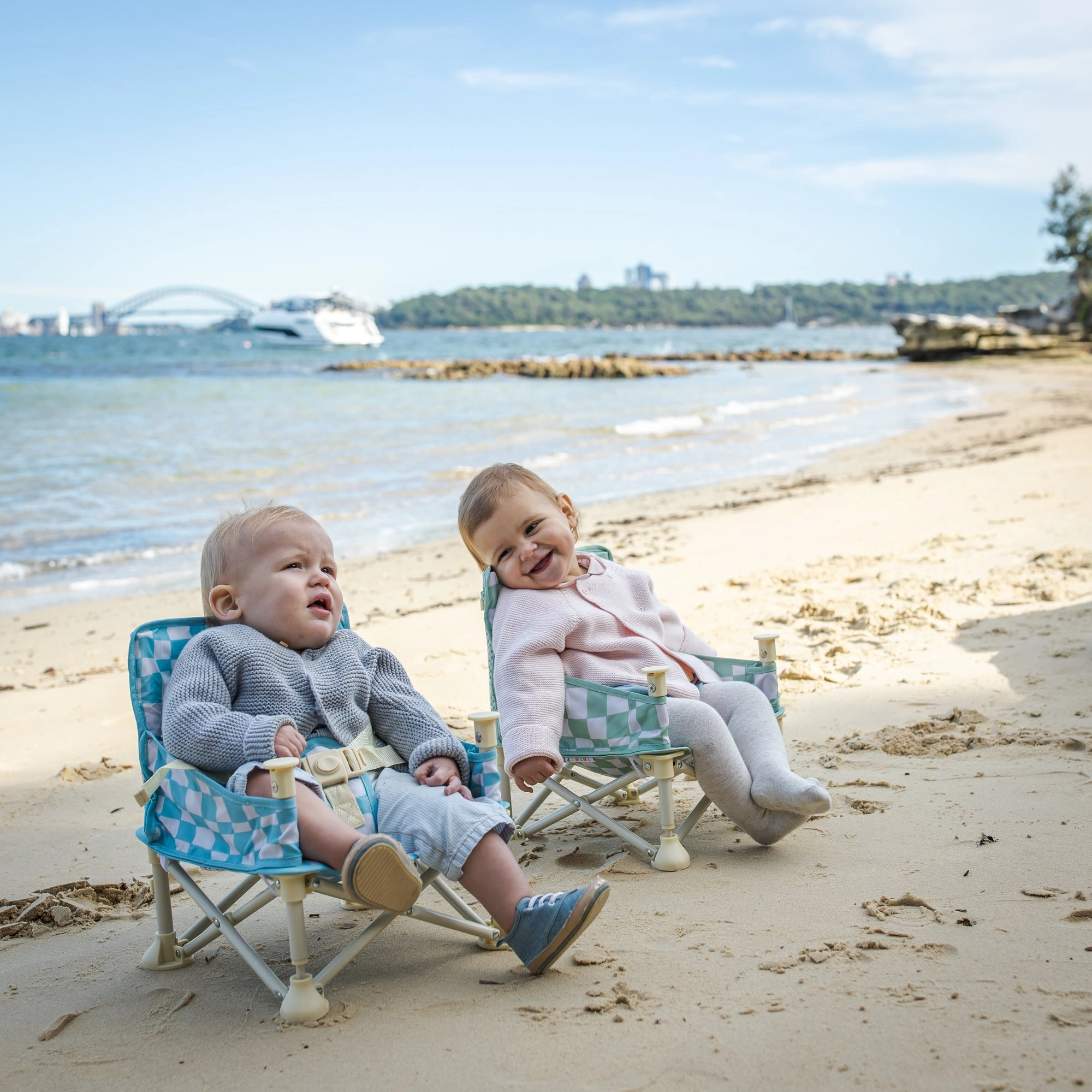 Two babies sitting side by side on the sand near the harbour