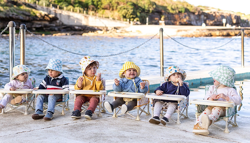 Six babies in a row sitting in beach chairs by the ocean