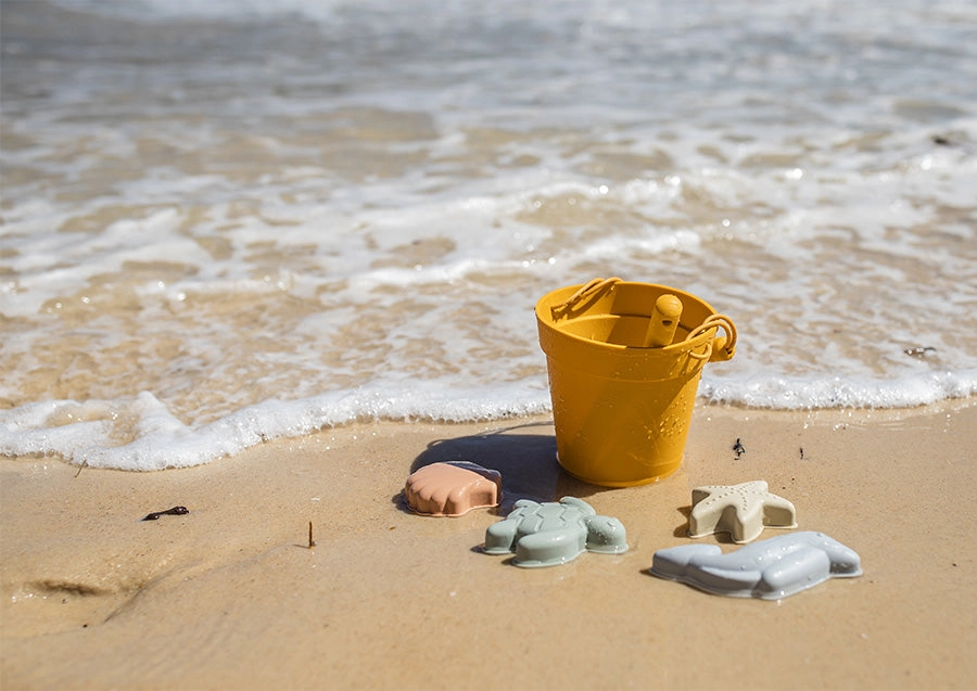 Toy bucket and beach shells in the sand as a wave washes over them